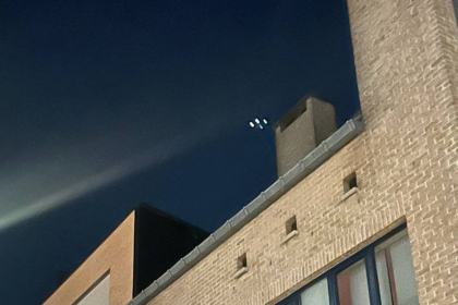 Resident night-mode photo of a light (with motion blur) above a lit building, published as one of "two drones". 2025-11-11 (evening), Between Heusden-Zolder and Beringen, Limburg, Belgium. Status: Police went to the scene after reports; nothing confirmed. Note: Metabunk: very likely an airliner (TUI and Brussels Airlines flights on matching tracks); long-exposure blur.